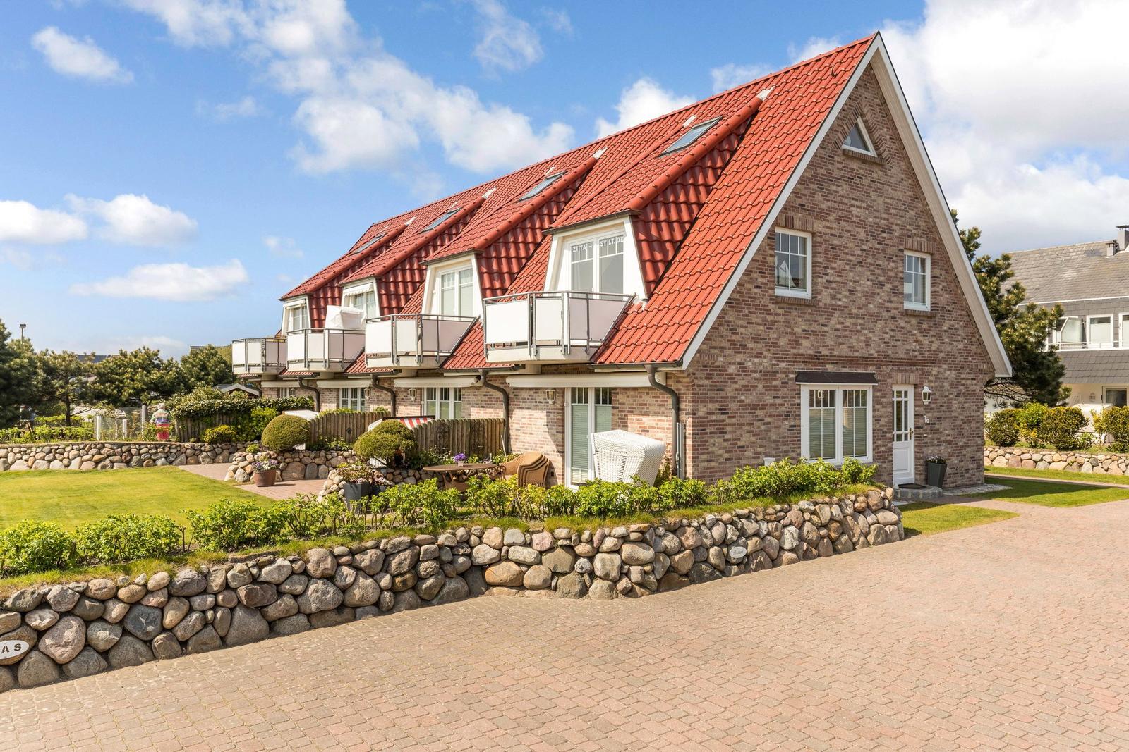 Ferienhaus mit rotem Dach und Balkonen, umgeben von Grünflächen und Steinmauern.