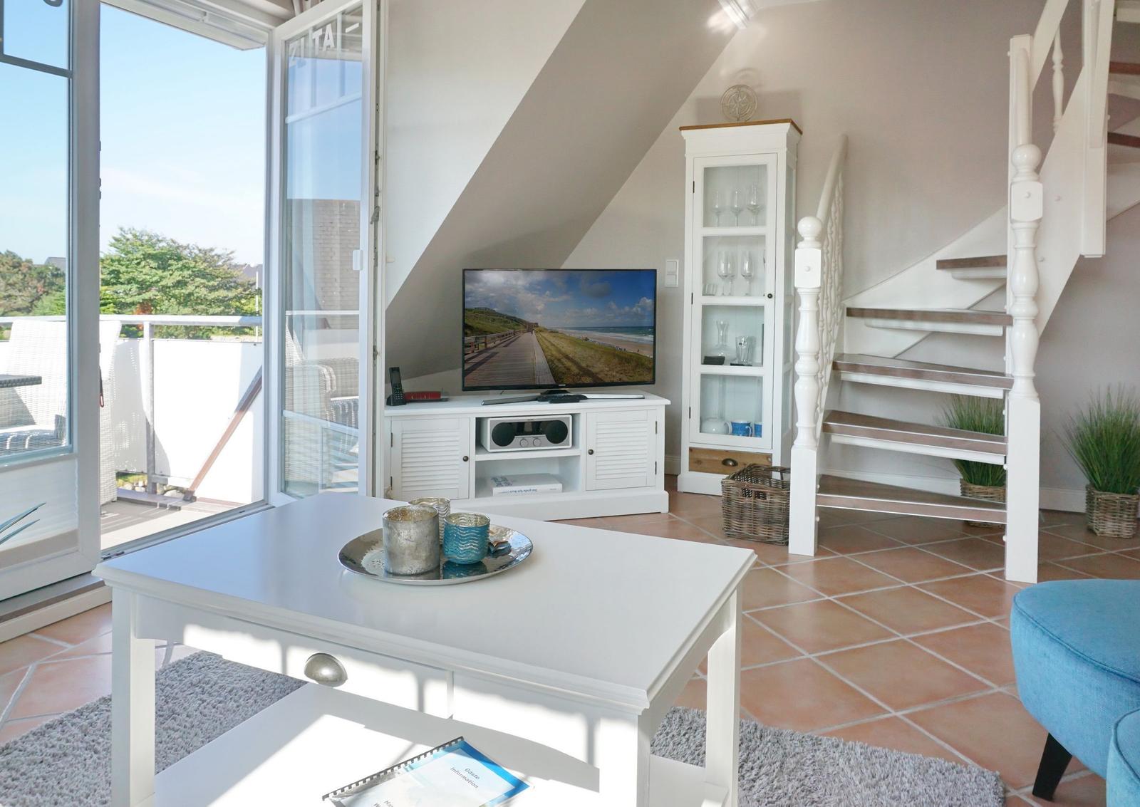 Wohnzimmer mit TV, Glasvitrine und Terrasse. Holztreppe und Terrassenmöbel sichtbar.
