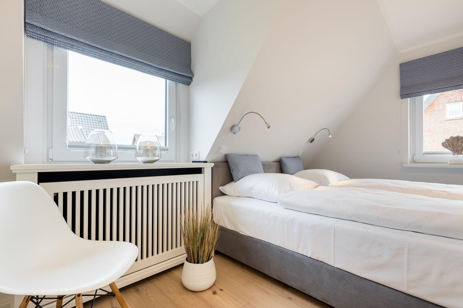 Doppelbett mit weißen Laken, zwei Fenster mit Rollladen und weißer Stuhl.