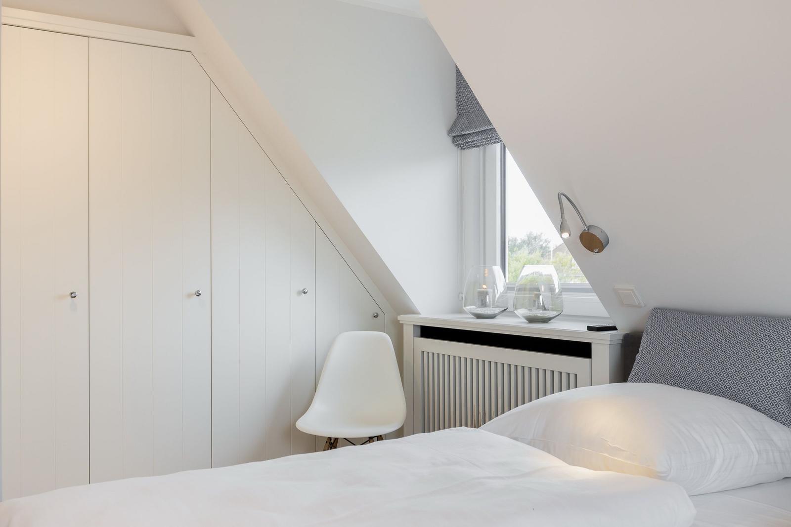 Schlafzimmer mit weißem Bett, Schrank und Fenster unter Dachschräge.