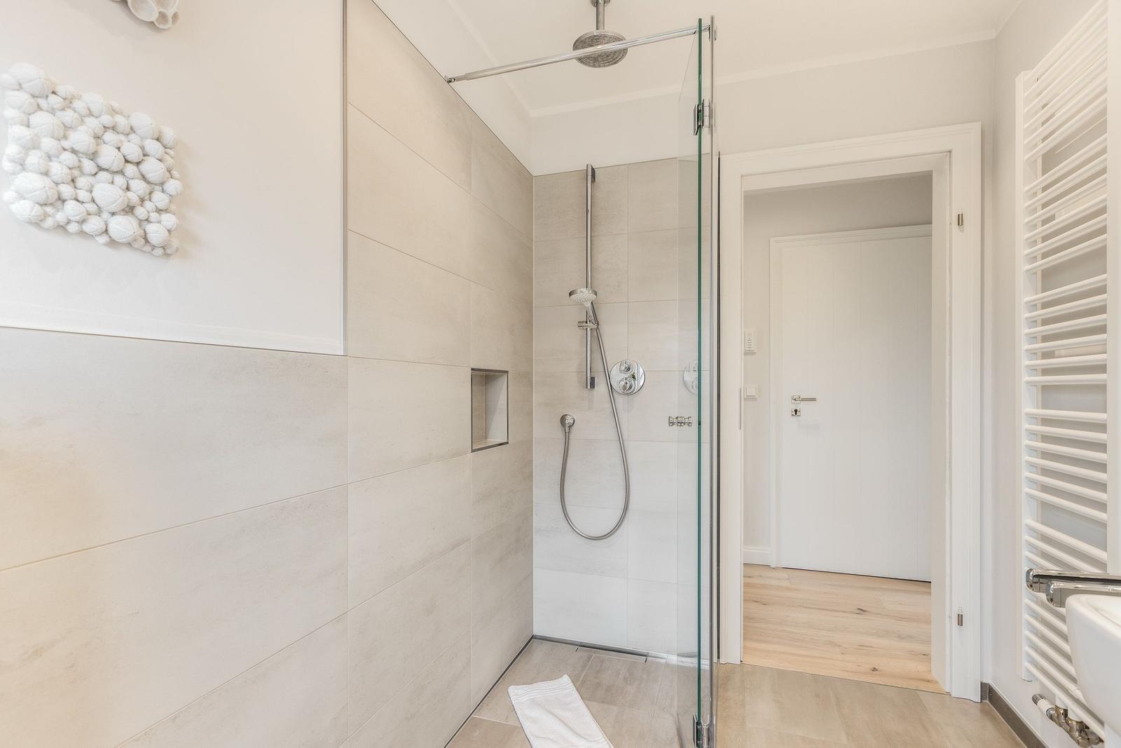 Dusche mit Glaswand, Duschbrause und Handbrause. Wanddekoration und Heizkörper sichtbar.