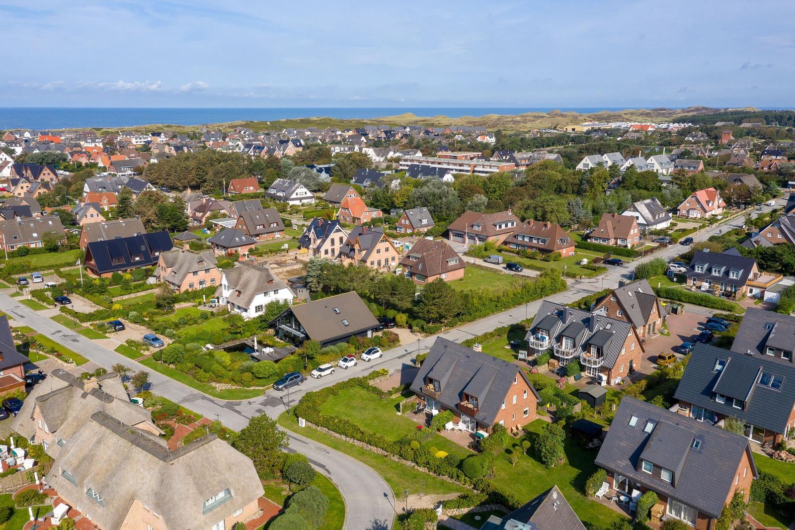 Aerial view of a coastal town with houses, greenery, and the sea in the background.