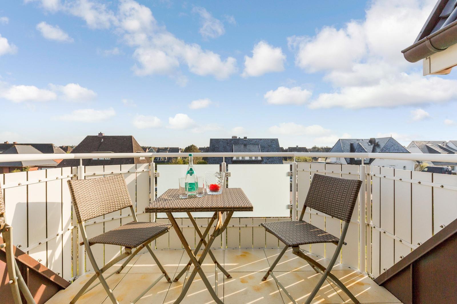 Terrasse mit Tisch und zwei Stühlen. Hintergrund: Dächer und blauer Himmel mit Wolken.