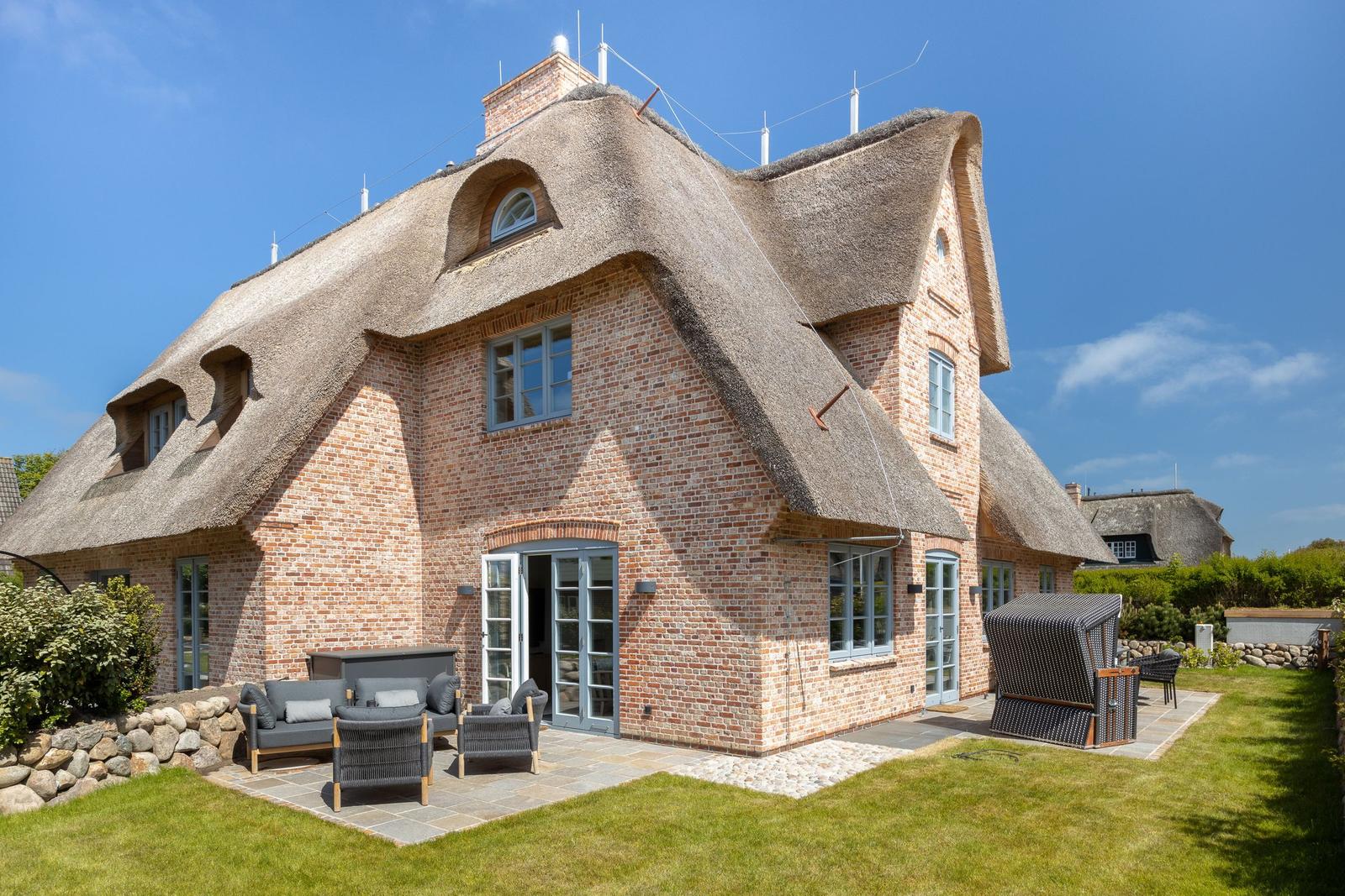House with thatched roof and stone wall. Terrace with seating and lawn area.
