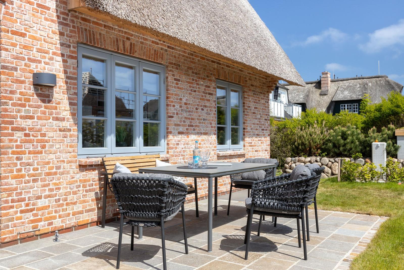 Terrace with table and chairs in front of a brick house with thatched roof.