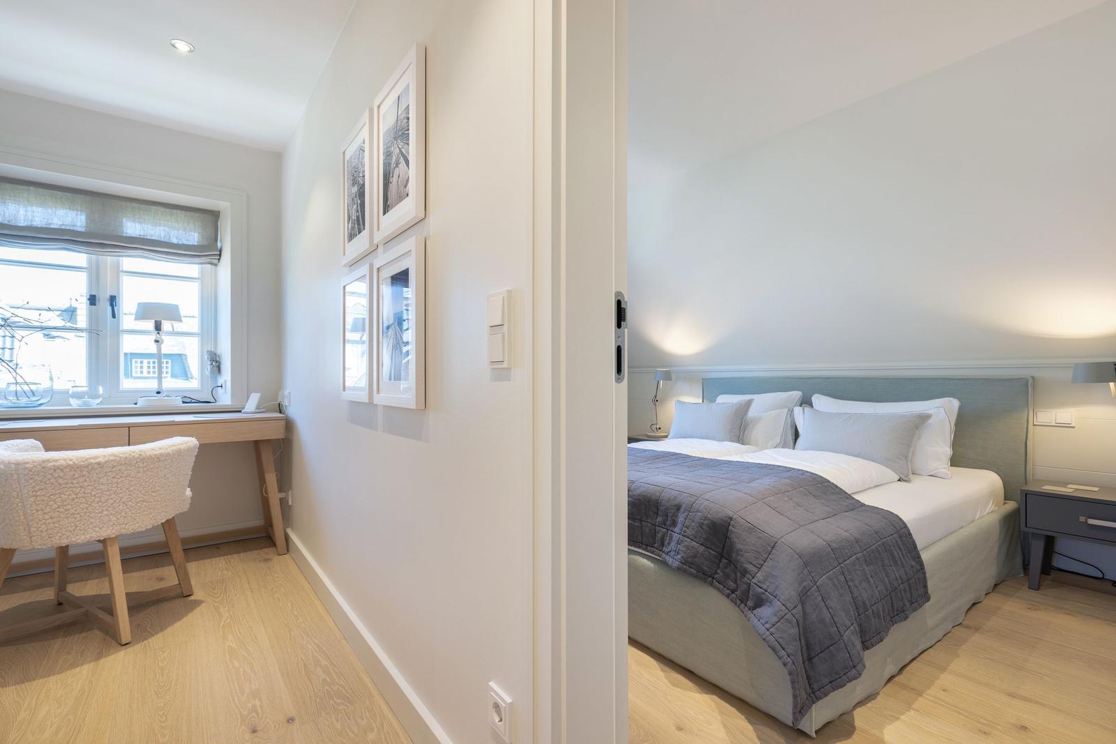 Bedroom with bed and nightstand. Adjacent room with desk and window.