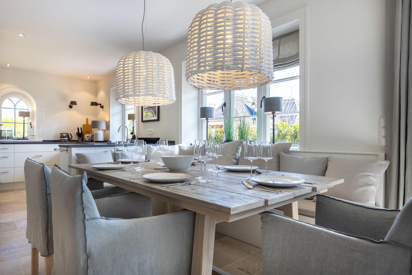 Dining area with wooden table, chairs, and large windows. Pendant lights and kitchen area in background.