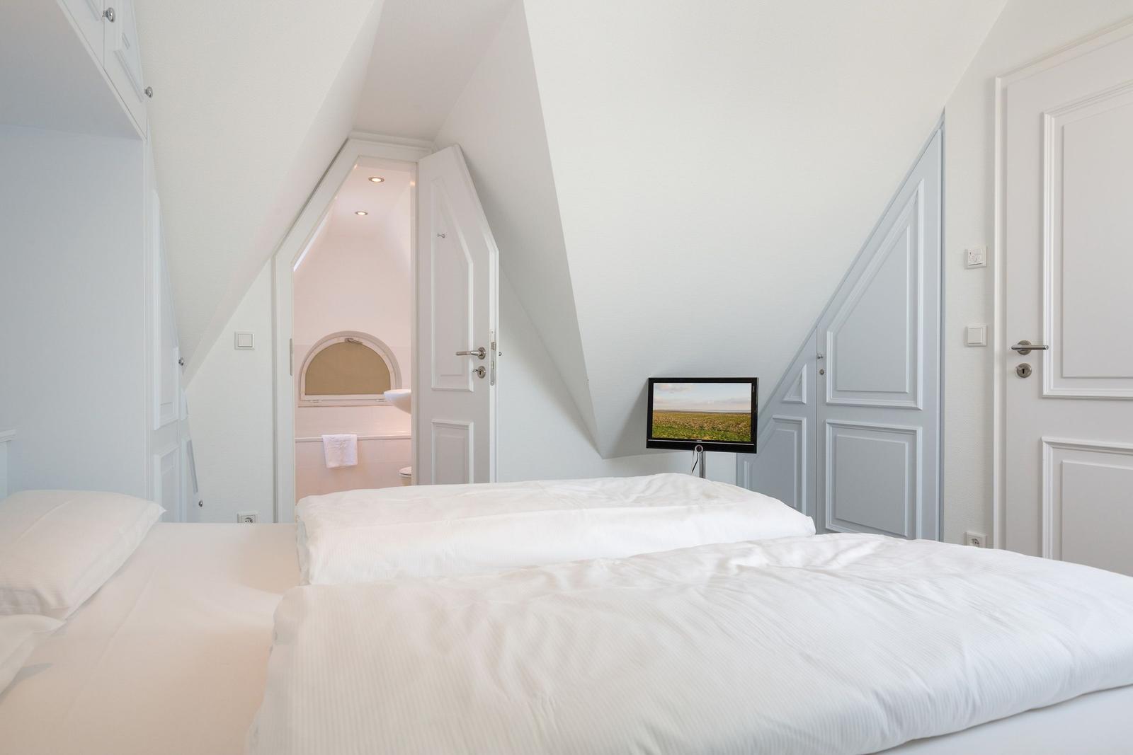 Schlafzimmer mit Dachschrägen, Bett und TV. Tür führt in Bad mit Rundfenster.