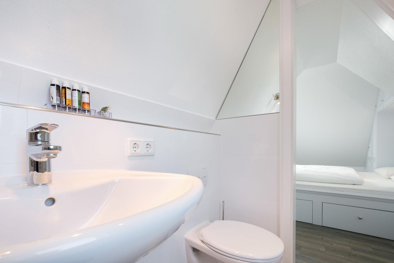 Badezimmer mit Waschbecken, Spiegel und Toilette. Blick in Schlafzimmer mit Bett.