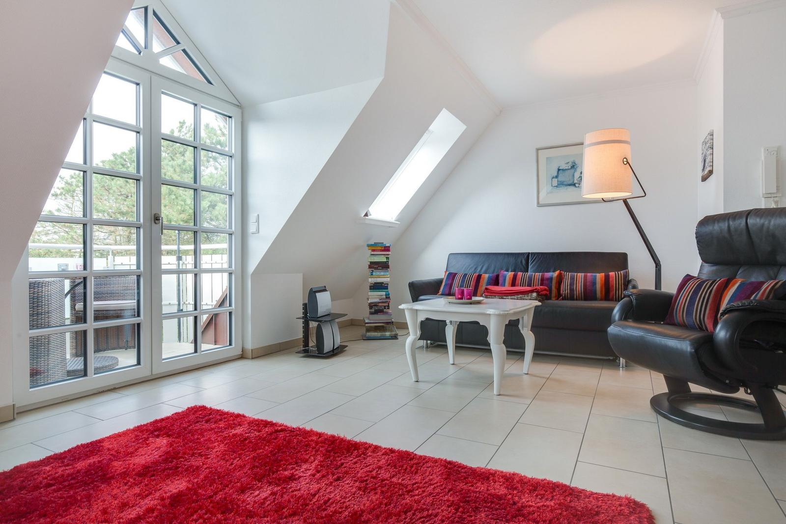 Wohnzimmer mit Sofa, Sessel, Tisch und rotem Teppich. Große Glastür führt auf Balkon.