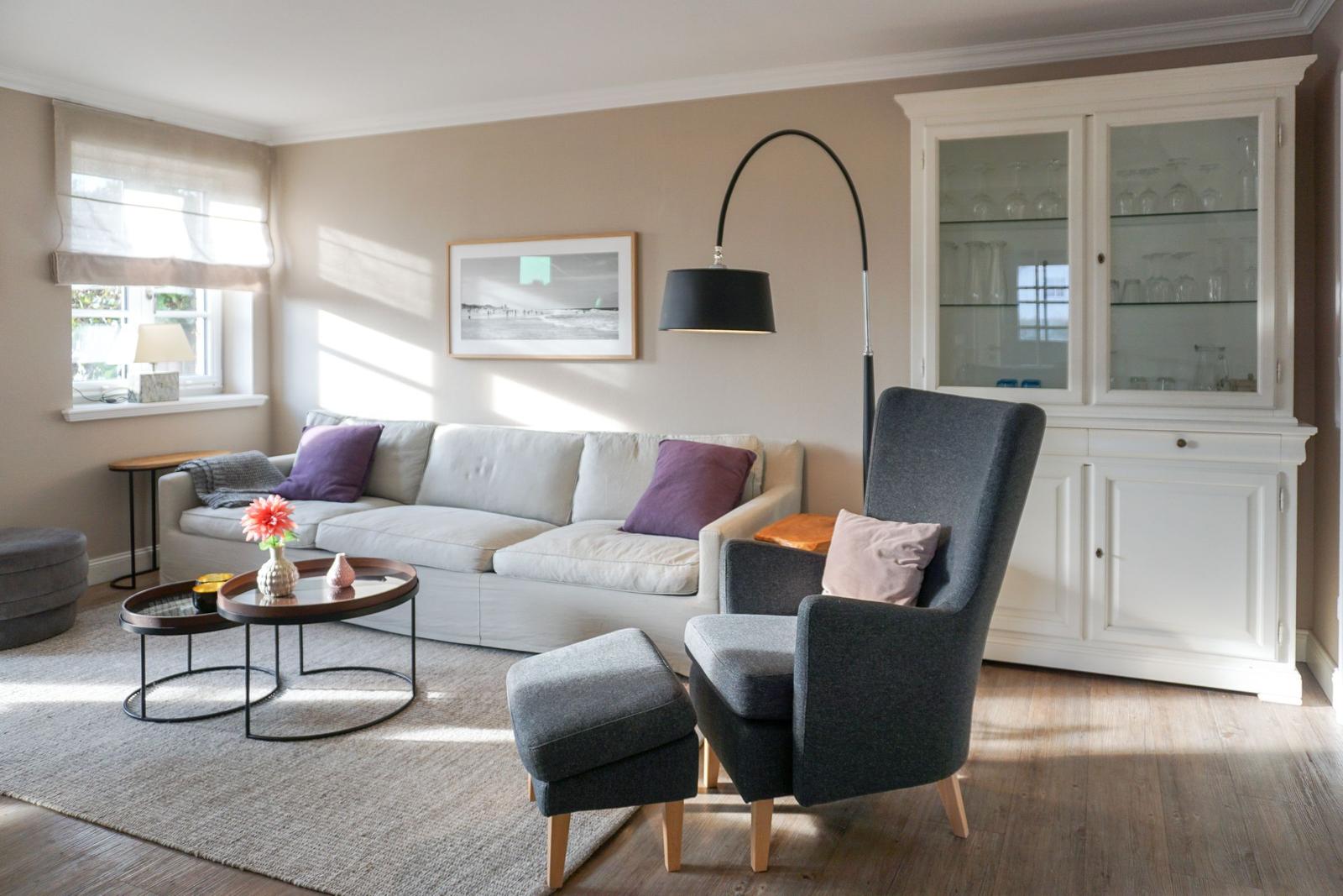 Living room with sofa, armchair, and glass cabinet. Wooden floor and window with roller shades.