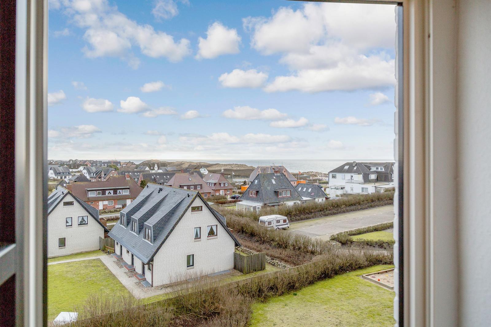 Aus dem Fenster sieht man Häuser und das Meer unter einem blauen Himmel mit weißen Wolken.