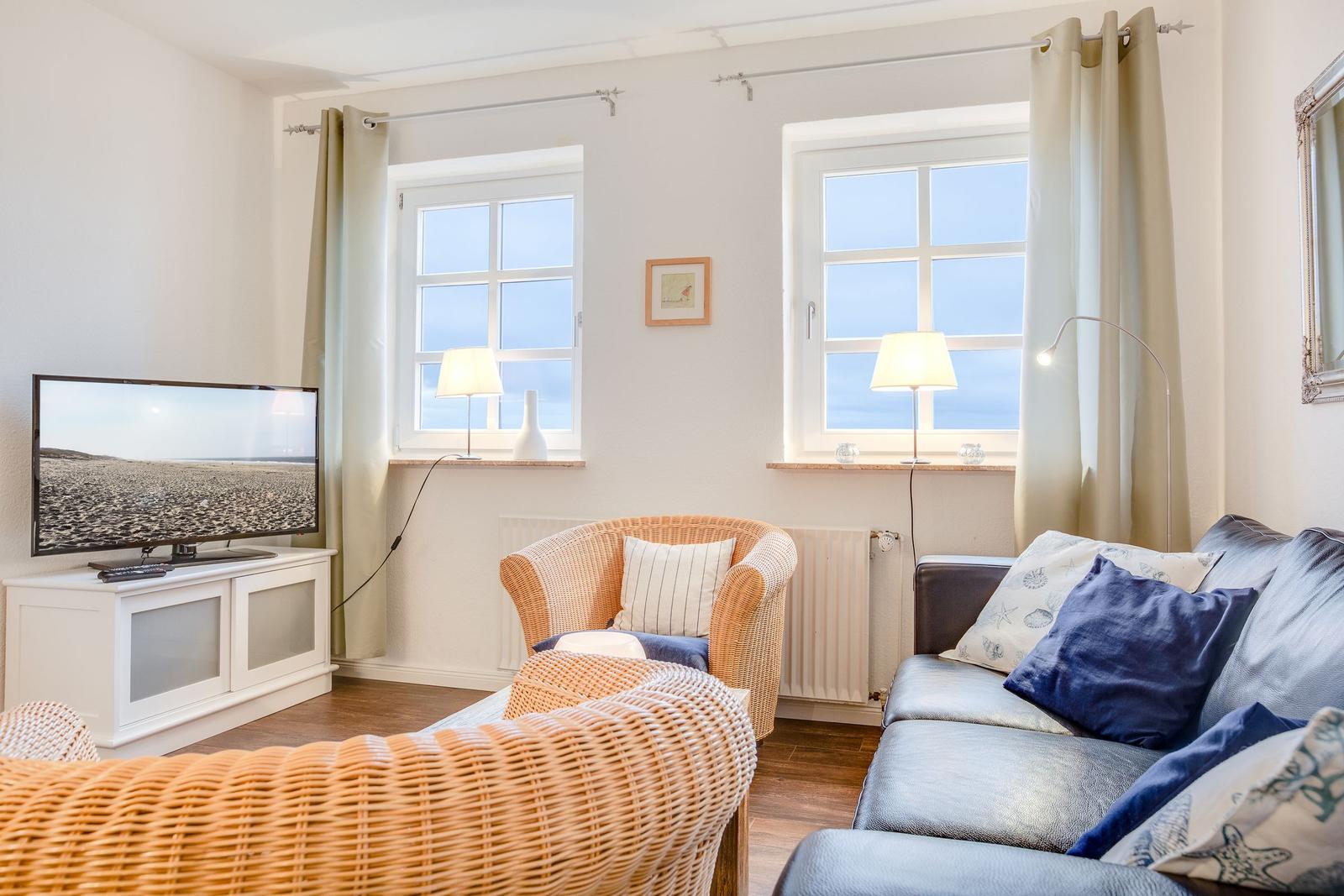 Wohnzimmer mit Sofa, Sessel, Fernseher und zwei Fenstern.
