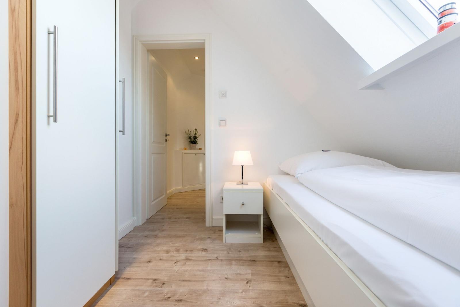 Bedroom with sloped ceiling, white bed and nightstand. Wooden floor and wardrobe.