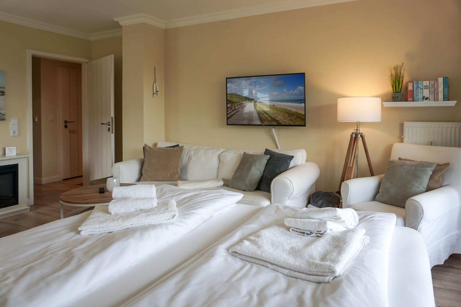 Two beds with towels, sofa, TV, and floor lamp in a bright room.