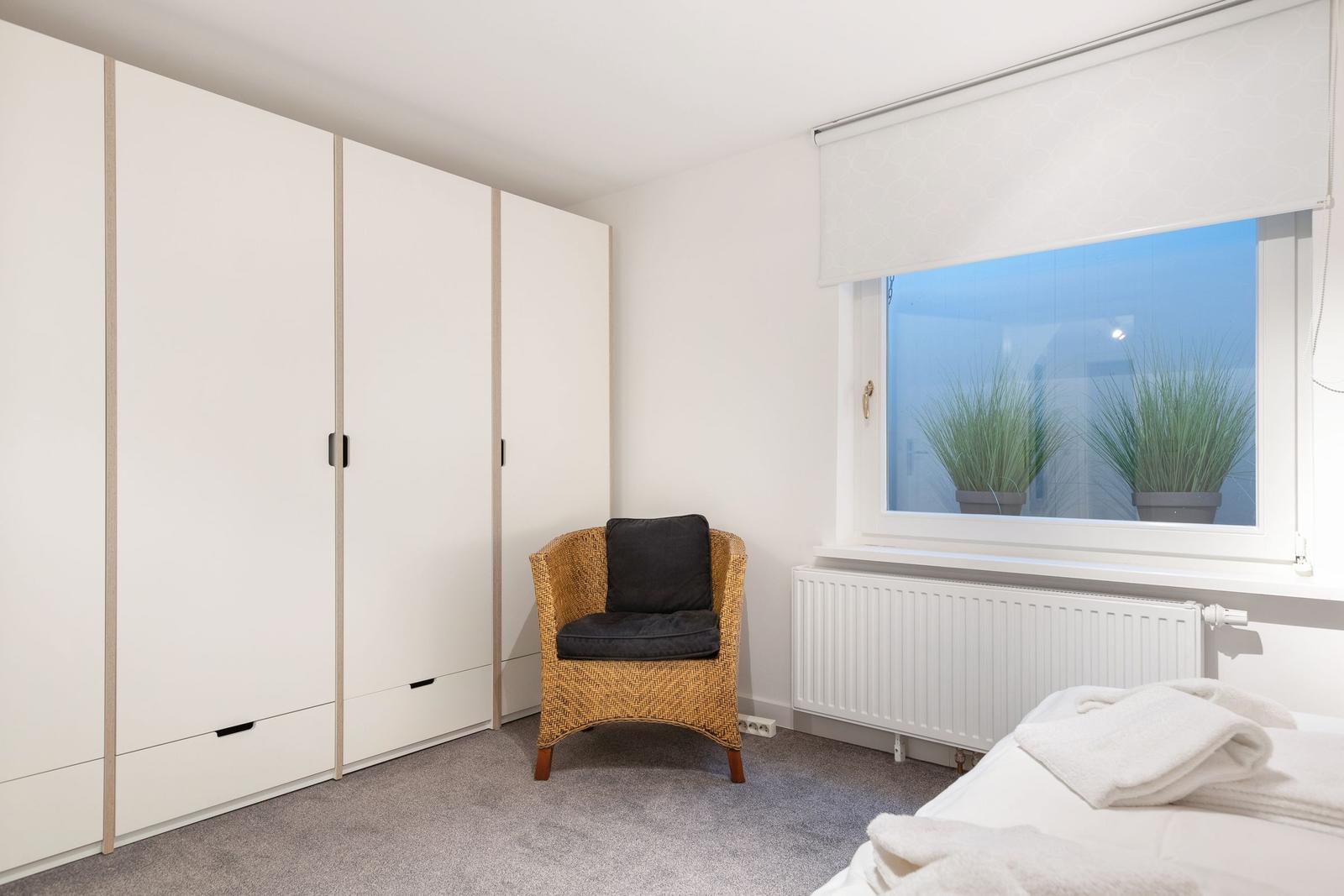 Schlafzimmer mit Bett, Kleiderschrank und Sessel vor Fenster.