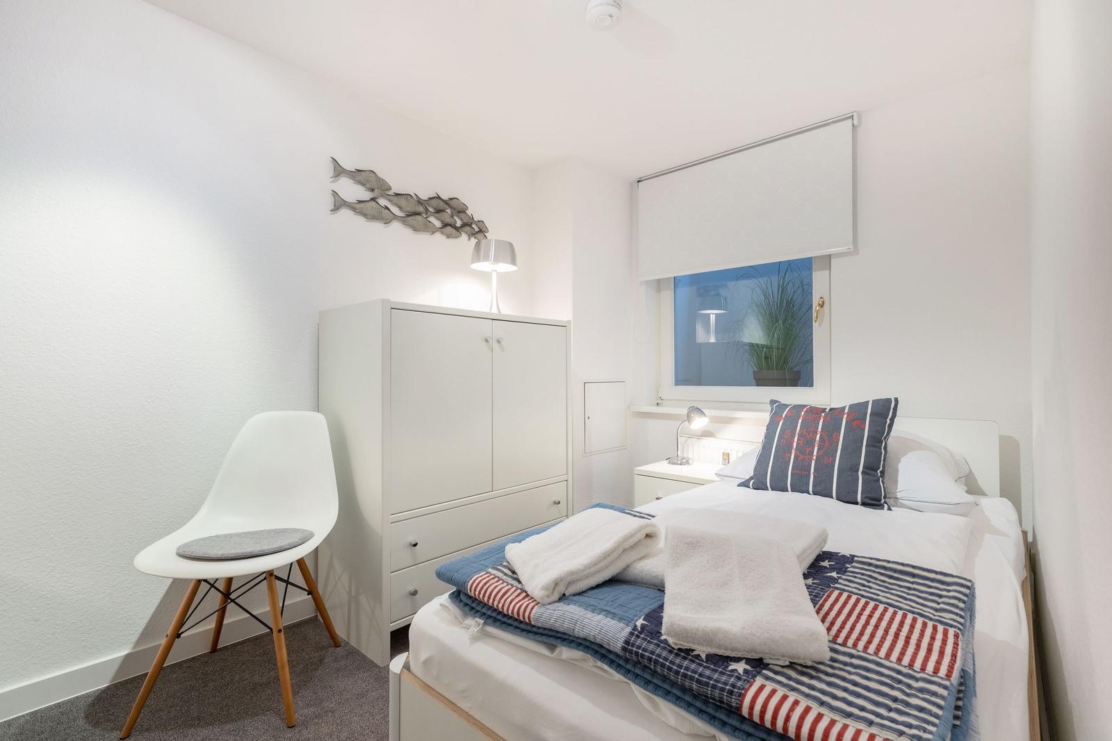 Schlafzimmer mit Bett, Kleiderschrank und Stuhl. Fenster mit Rollladen und Pflanze.