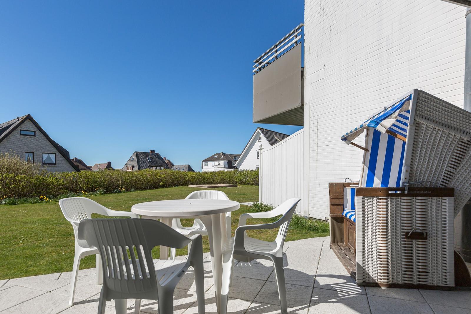 Terrasse mit weißem Tisch und Stühlen, Strandkorb und Blick auf Grünfläche und Häuser.