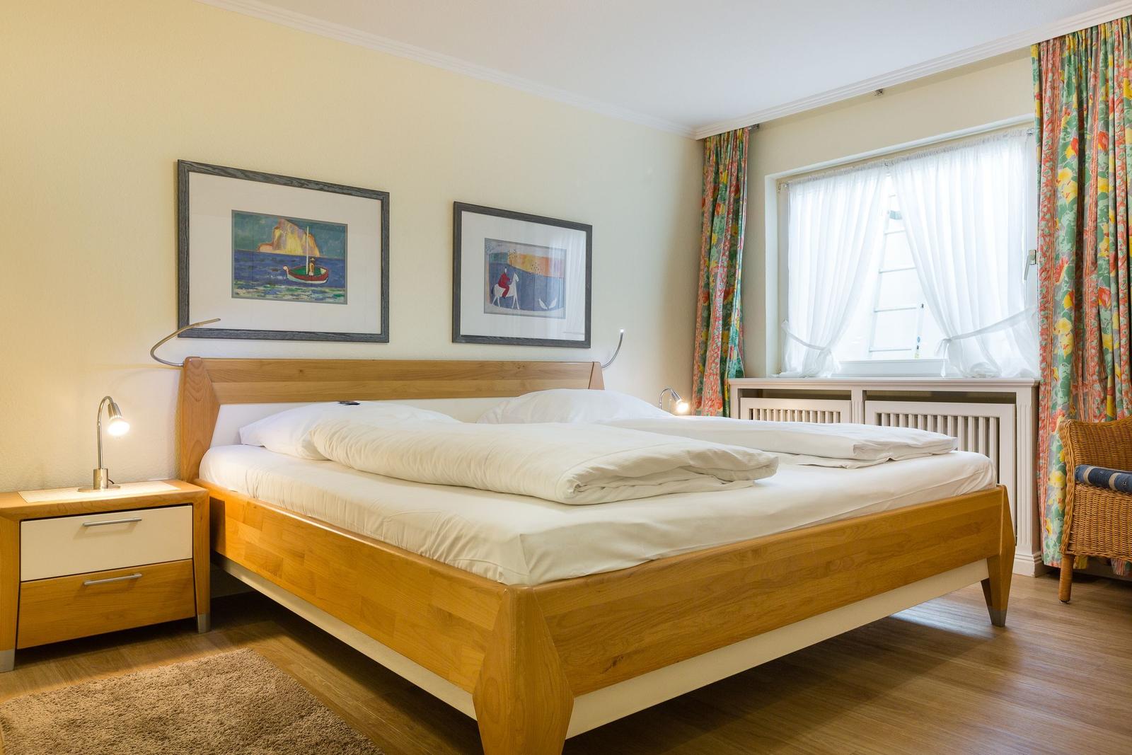 Schlafzimmer mit Holzbett, Nachttisch und Fenster mit Vorhängen.