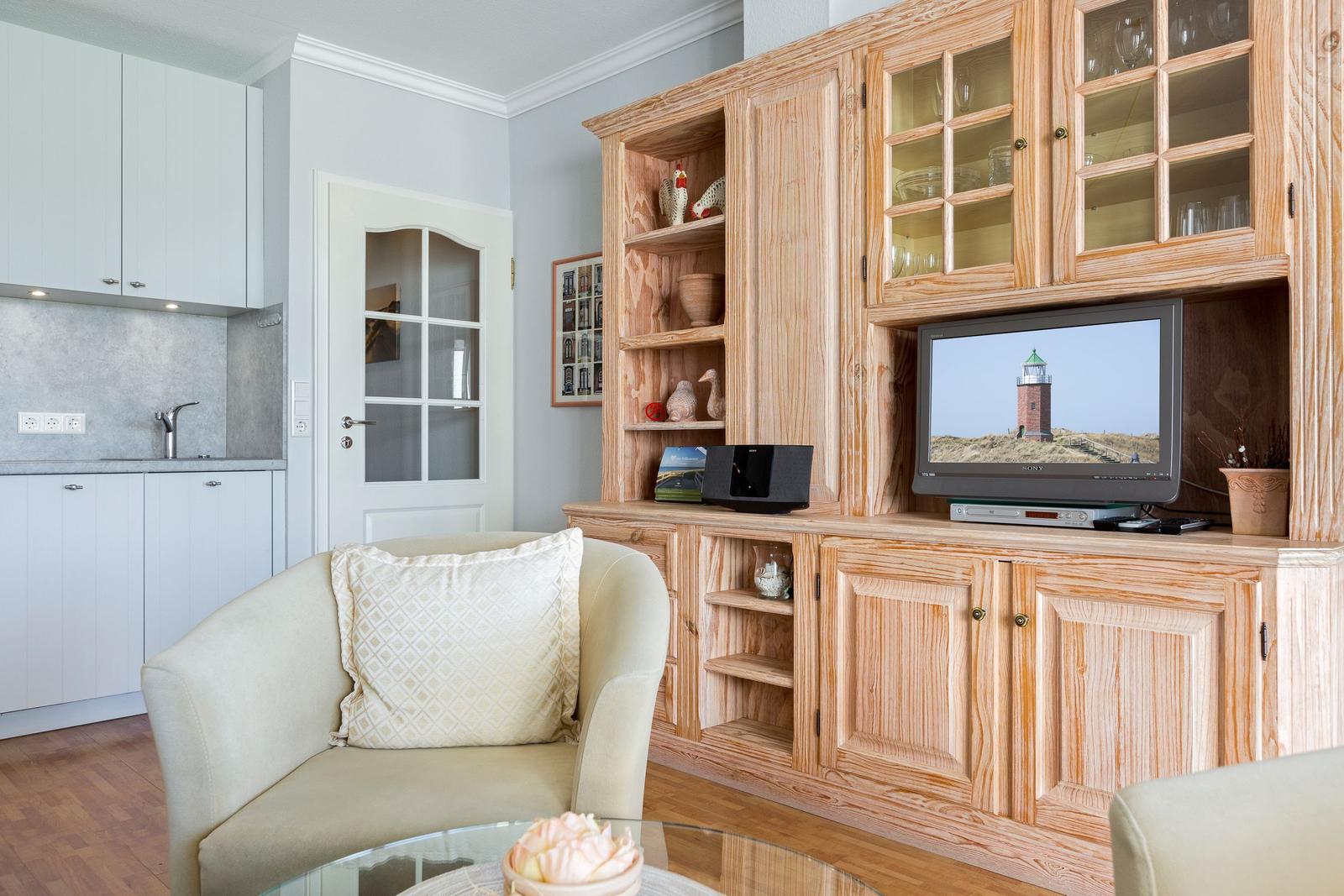 Wohnzimmer mit Holzregal, Fernseher und Sitzgelegenheit. Küche im Hintergrund.