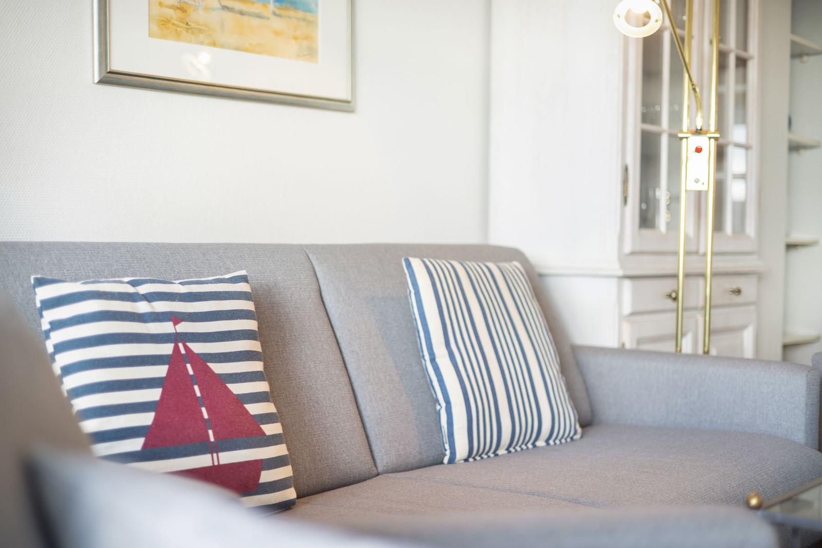 Graße Couch mit Kissen, eines mit Segelbootmotiv, vor weißer Wand und goldener Stehlampe.