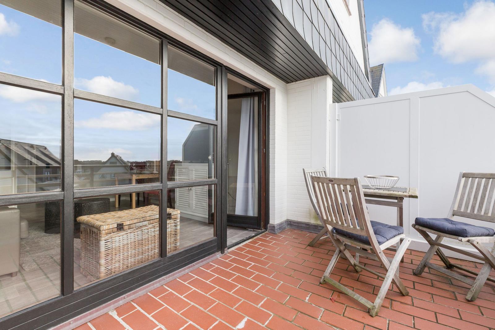 Terrace with wooden chairs and table, red tiles, and glass wall to interior.