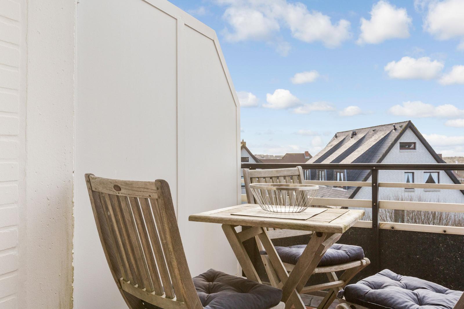 Terrace with wooden table and two chairs with cushions. View of rooftops under blue sky.