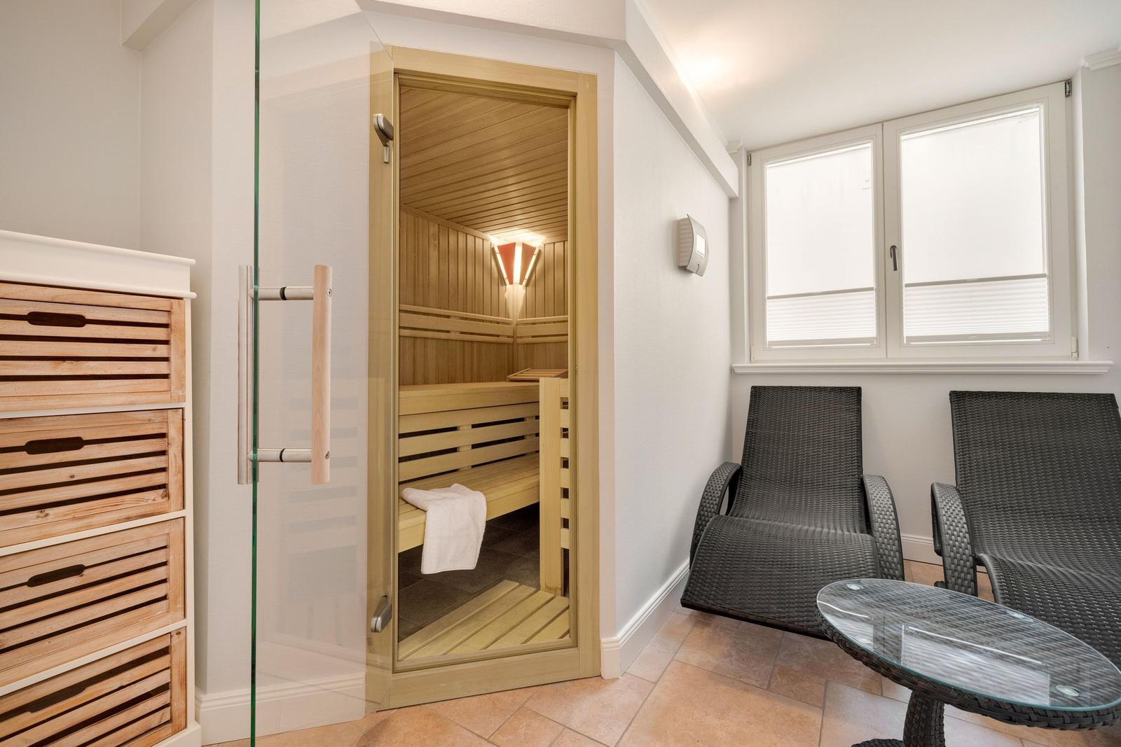 Sauna with wooden bench and glass door. Adjacent area with lounge chairs and table.