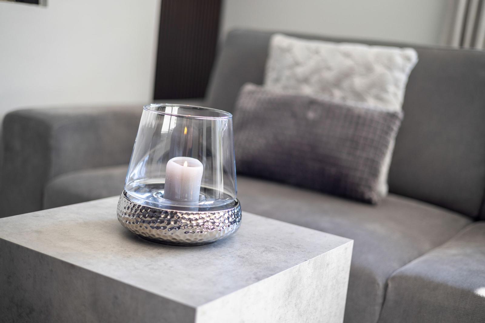 A lit candle in a glass on a table in front of a sofa with cushions.