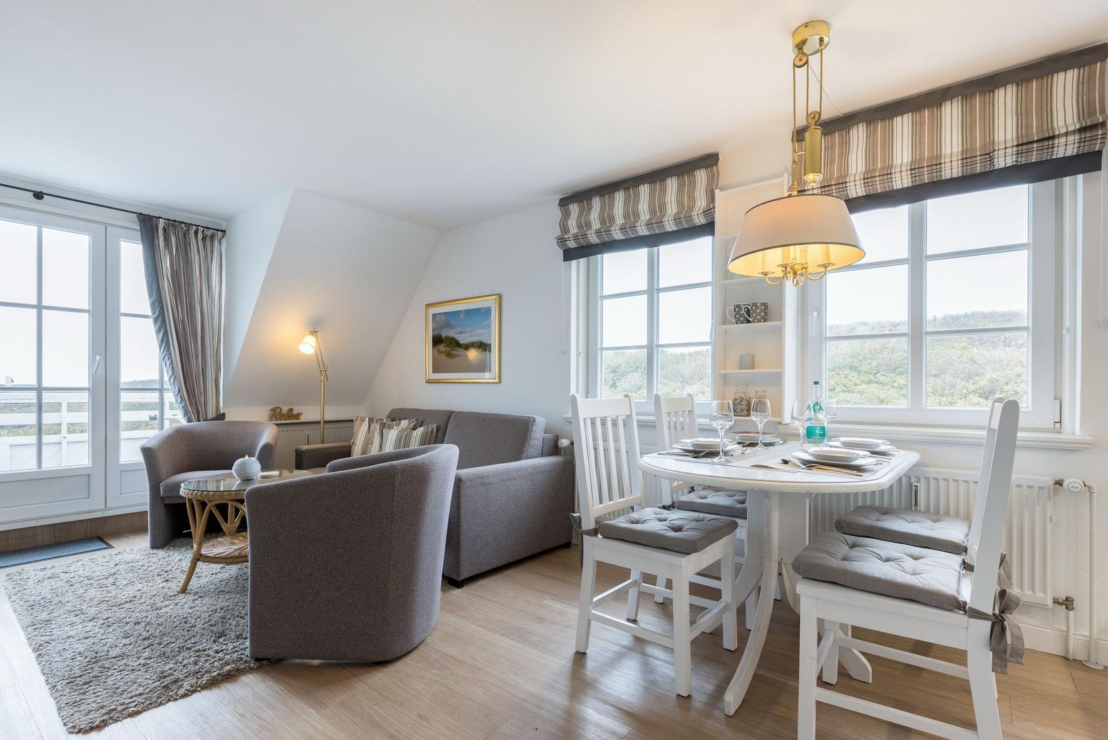 Living and dining area with sofa, chairs, and table. Large windows with view of landscape.