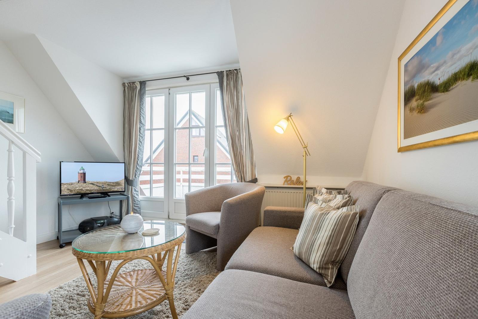 Living room with sofa, armchair, glass table, and TV. Window with view of house.