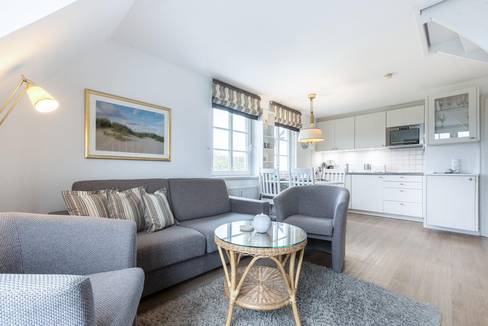 Living room with sofa, armchair, and kitchen area. Wooden floor and large windows.