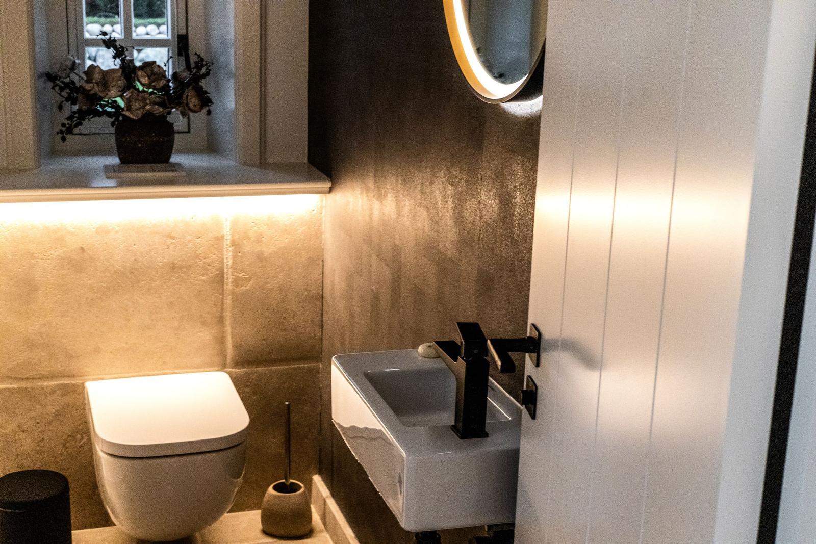 Badezimmer mit Toilette, Waschbecken und Spiegel. Fenster mit Blumen auf Sims.