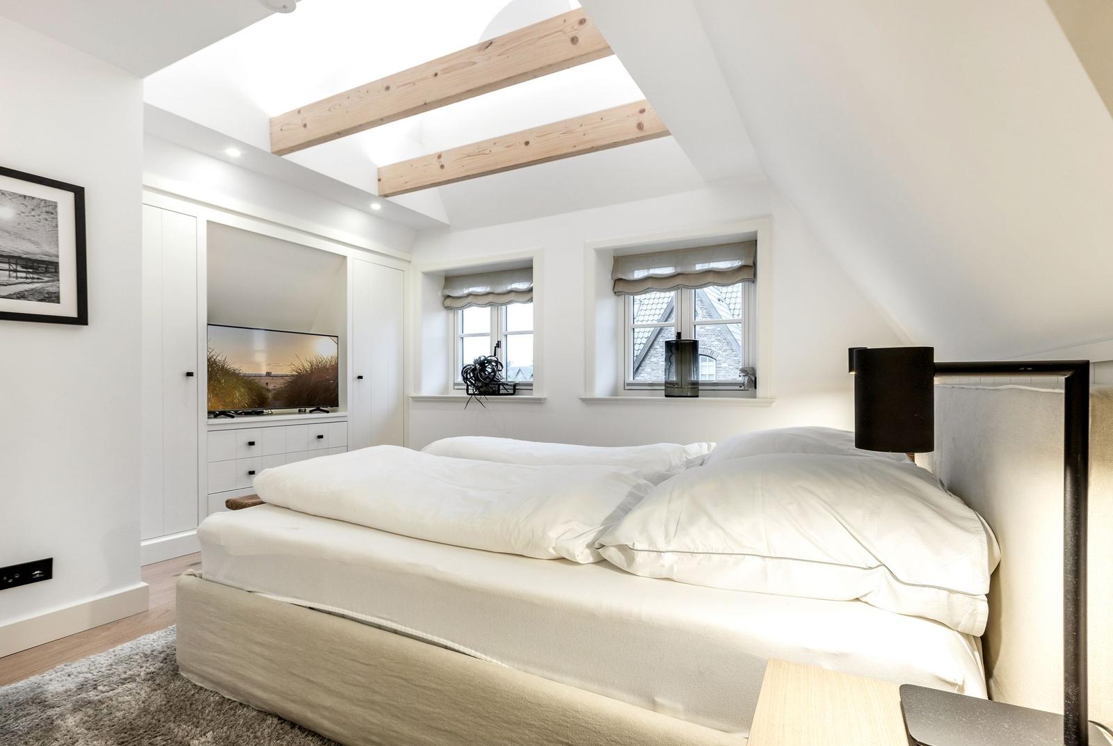 Schlafzimmer mit Dachschrägen, zwei Fenstern und einem Bett mit weißen Kissen.
