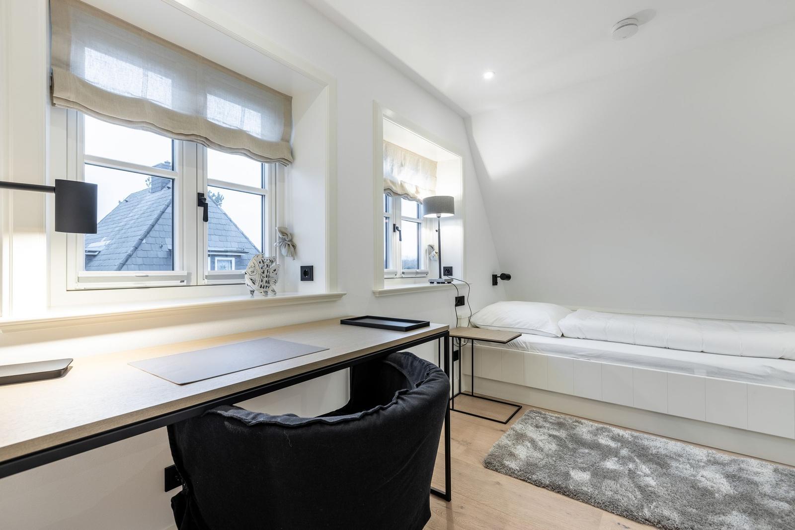 Schlafzimmer mit Schreibtisch, Bett und zwei Fenstern.