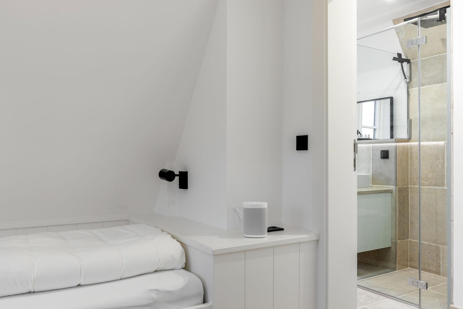 Schlafzimmer mit weißem Bett und integriertem Schrank. Nebenraum mit Dusche und Spiegel.