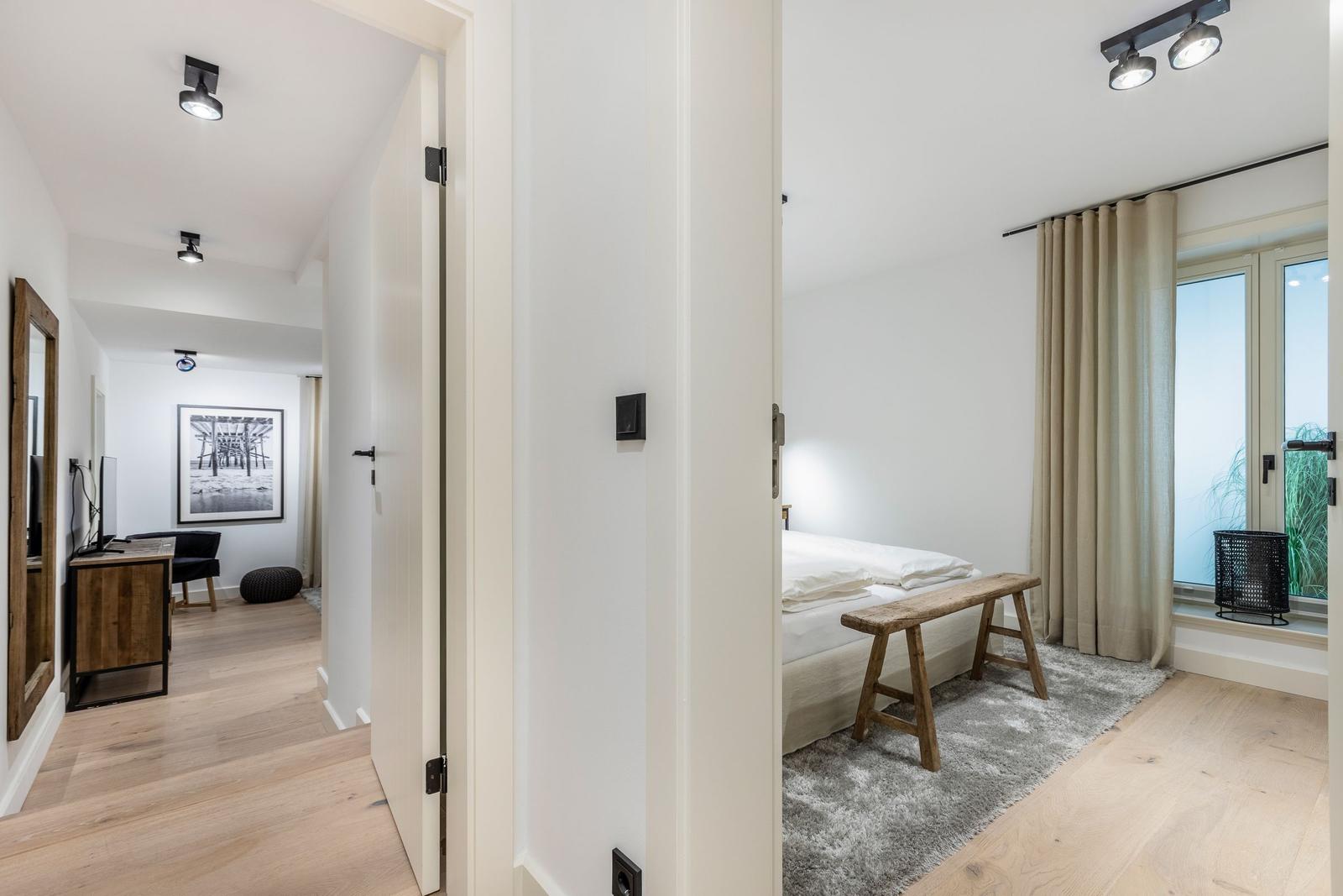 Schlafzimmer mit Bett, Holzbank und Fenster. Flur mit Schreibtisch und Wandbild.