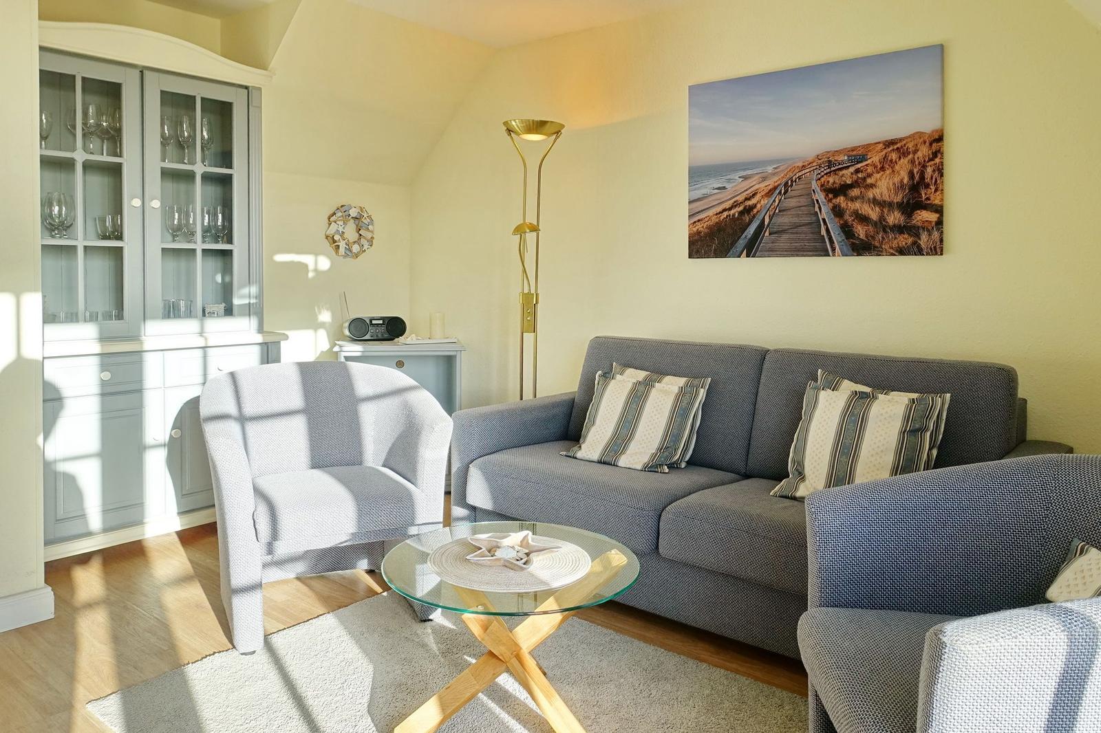 Living room with sofa, armchairs, and glass table. Wall art shows beach path.