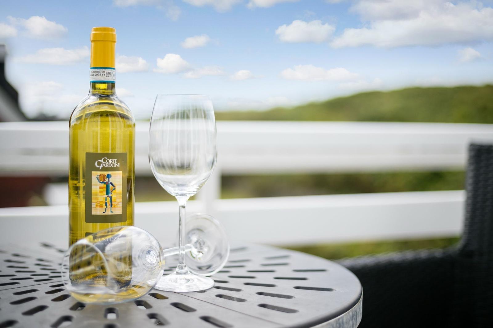 A bottle of white wine and two glasses sit on a terrace table with a view of green hills.