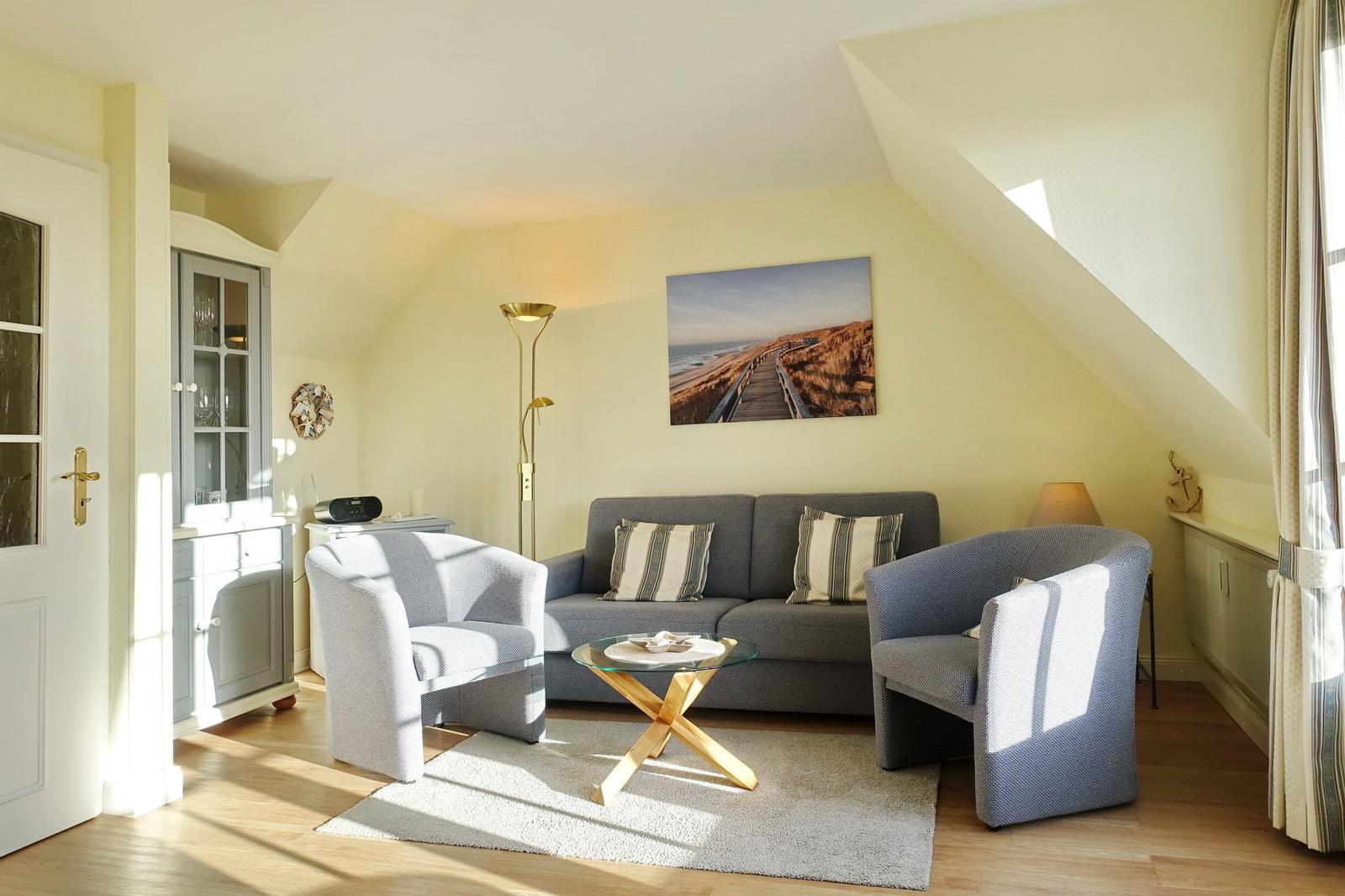Living room with sofa, two chairs, and glass table. Wall picture shows beach. Wooden floor and vaulted ceiling.