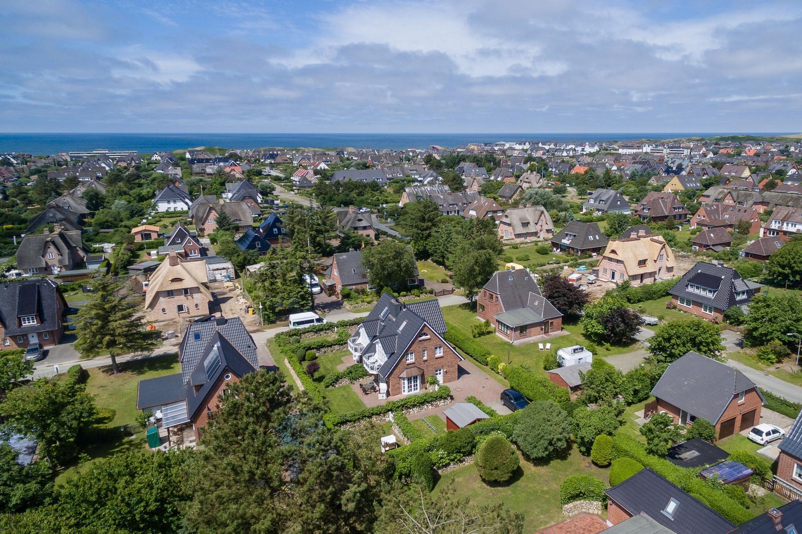 Aerial view of a coastal town with houses, greenery, and the sea in the background.