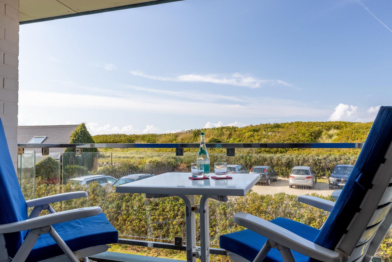 Balcony with two blue chairs and table. View over greenery and parked cars.