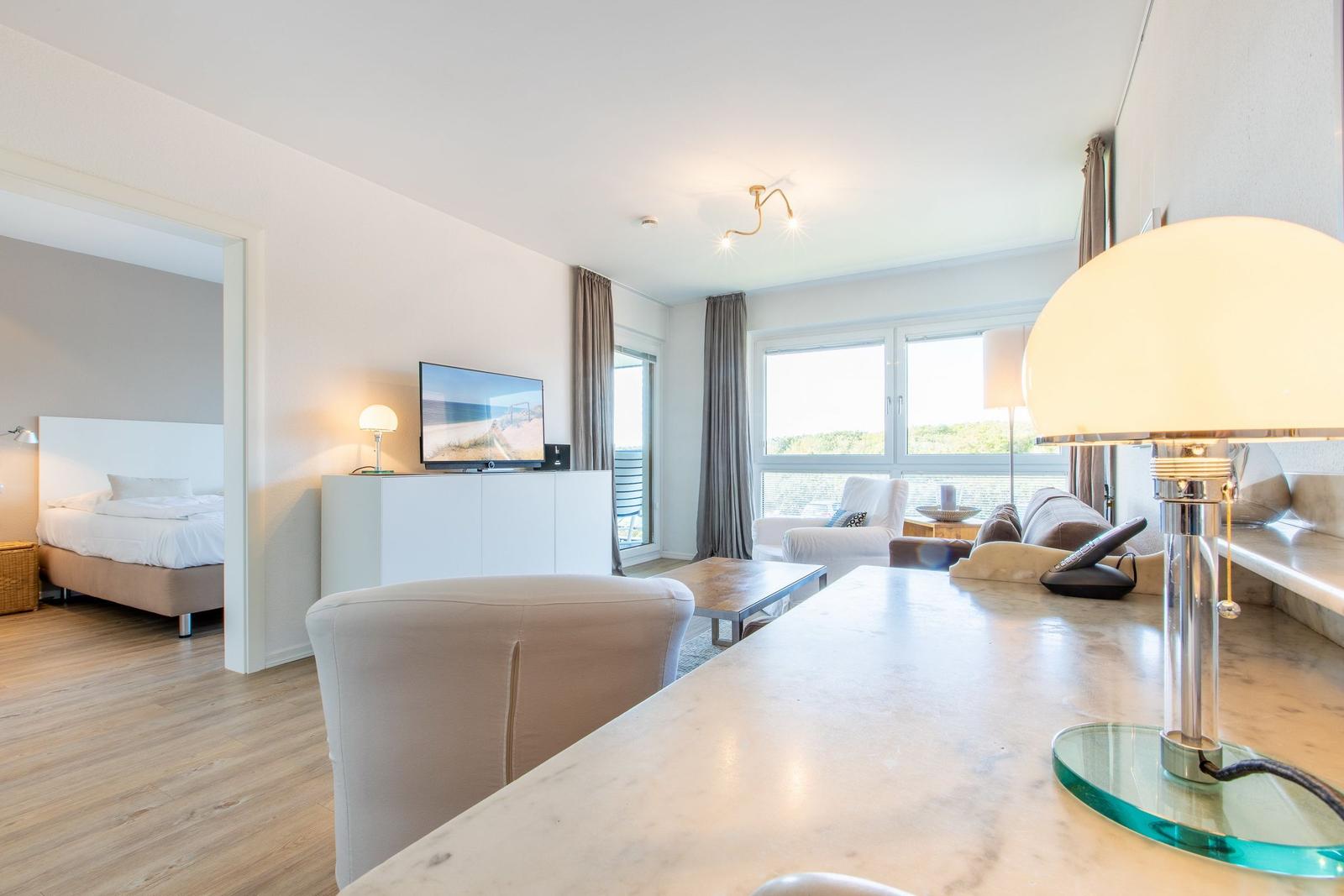 Living area with TV, sofa, and balcony view. Bedroom with bed in background.