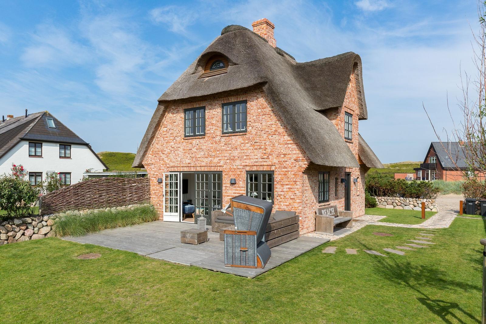 Red brick house with thatched roof and terrace with garden furniture.