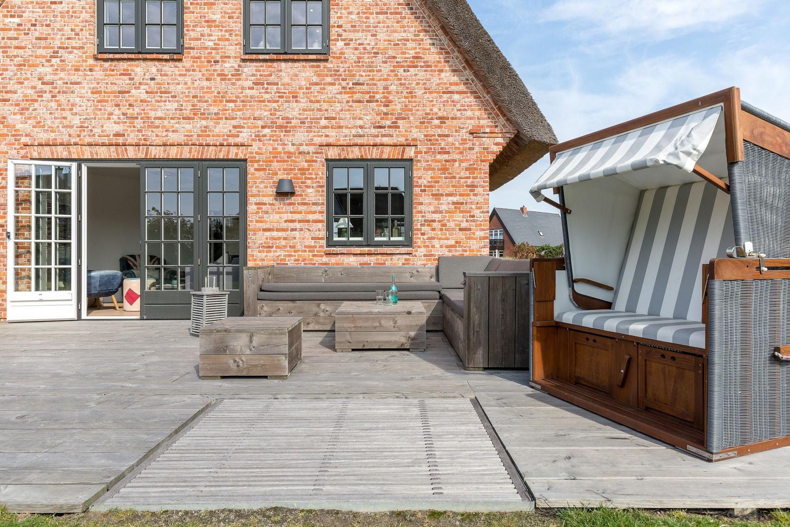 Terrace with wooden furniture and beach chair in front of brick house with sliding doors.