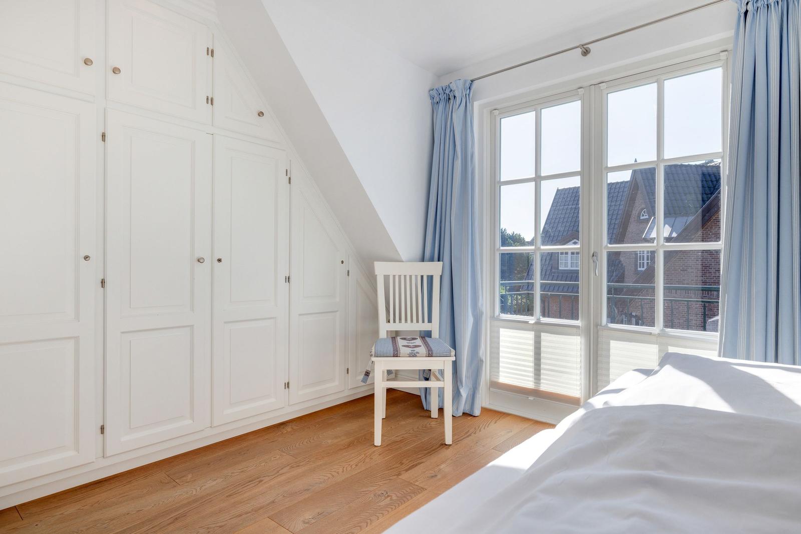 Schlafzimmer mit weißem Schrank, Stuhl und Fenster mit Blick auf Gebäude.