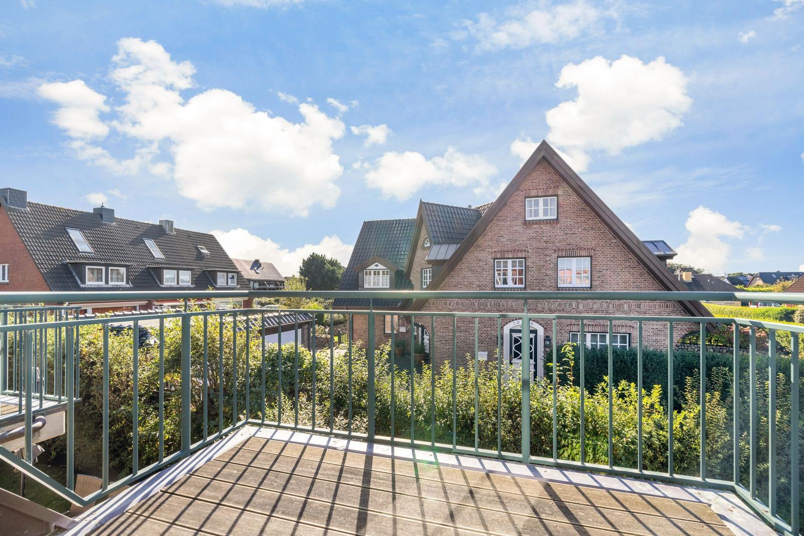 Blick von Balkon auf roten Backsteinhaus mit grünen Hecken und blauem Himmel.