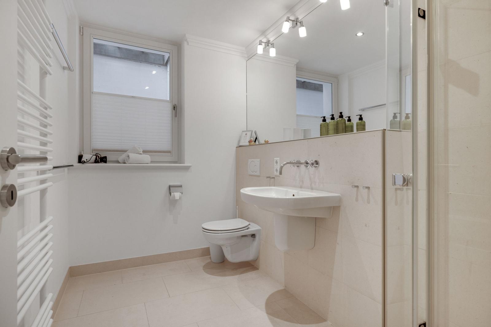 Badezimmer mit Toilette, Waschbecken und Dusche. Fenster mit Jalousie und Handtuchhalter.