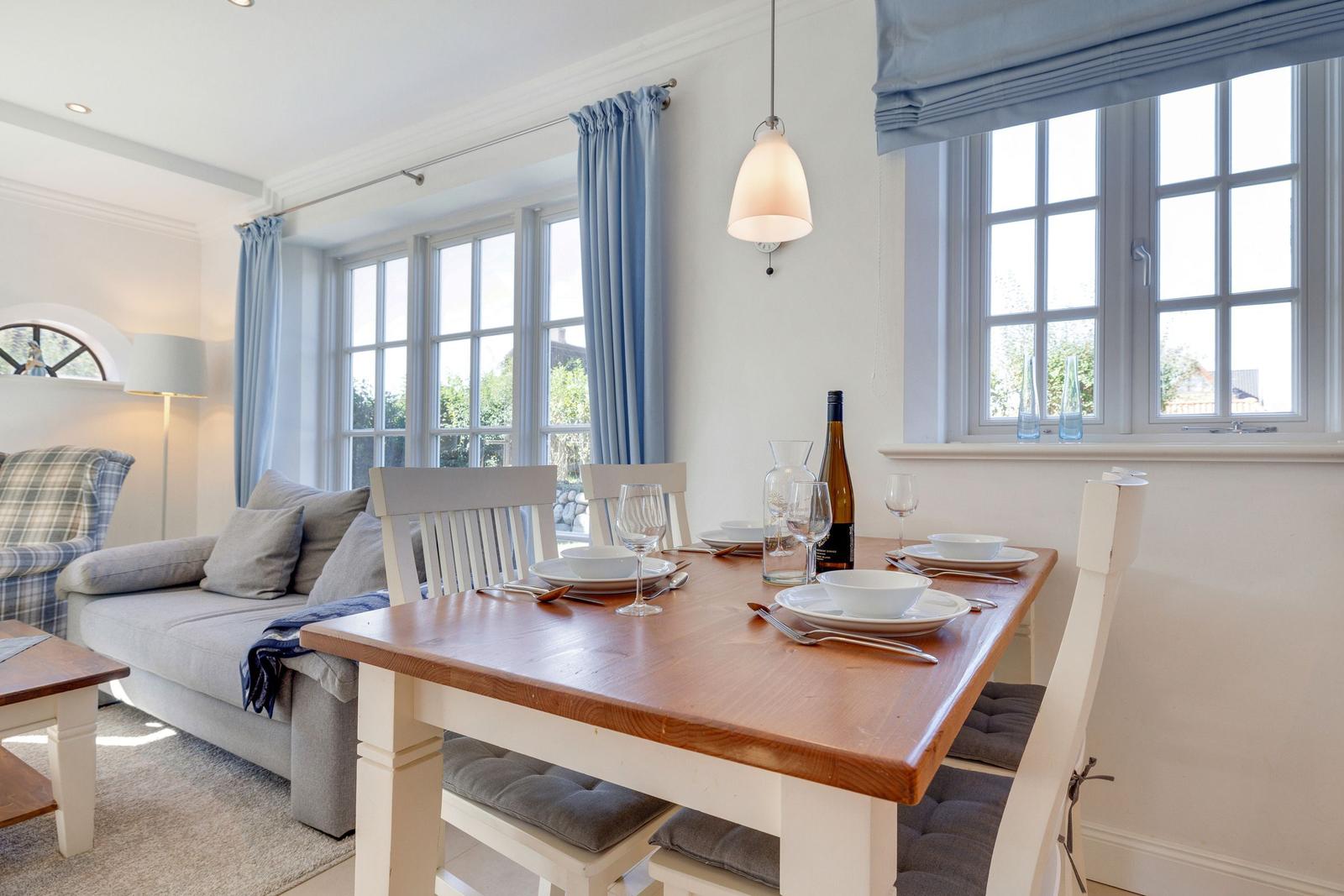 Esstisch mit Stühlen, Fenster mit Vorhängen und Sofa im Hintergrund.
