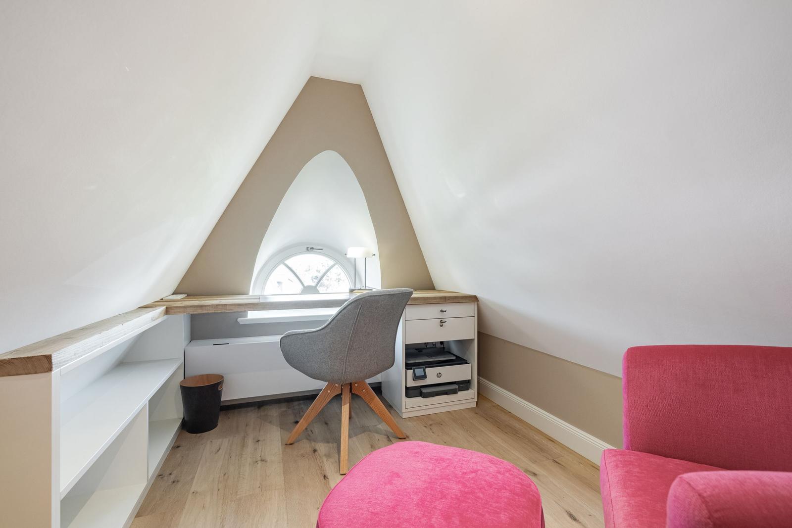 Dachgeschosszimmer mit Schreibtisch, rosa Sitzbank und Bogenfenster.